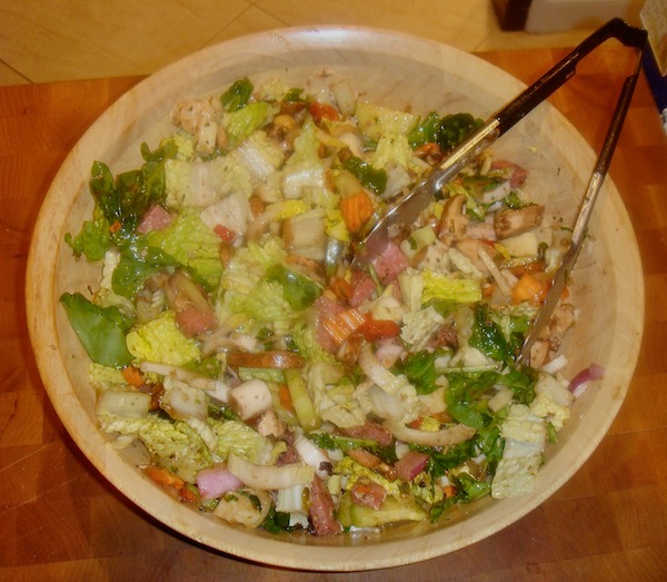 A large light wood salad bowl filled with greens, ham, cheese, tomatoes and carrots.