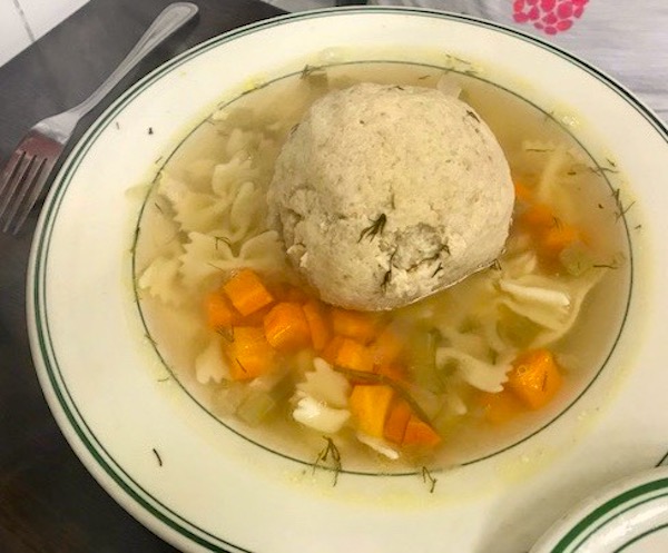 A white bowl with thin green stripes on the rim filled with chicken noodle soup with carrots and a large matzoh ball