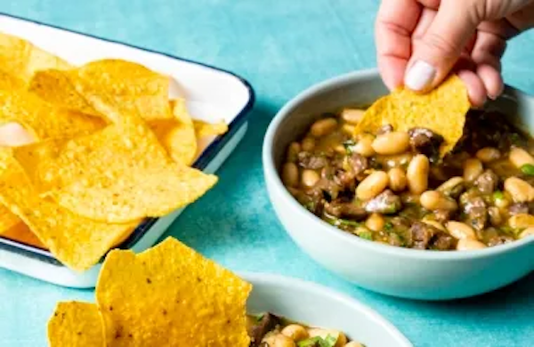 A white bowl filled with a bean and beef stew and a white pan with tortilla chips from A Trio from America's Test Kitchen