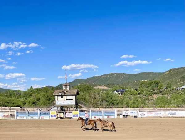 Rodeo set up with dirt on ground, broadcast stand fencing and 2 horses, one with rider and mountains in background at our Our First Rodeo Durango CO