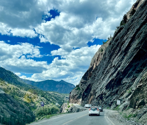 Dangerous mountain road with rock formations and shear drop visible on left.